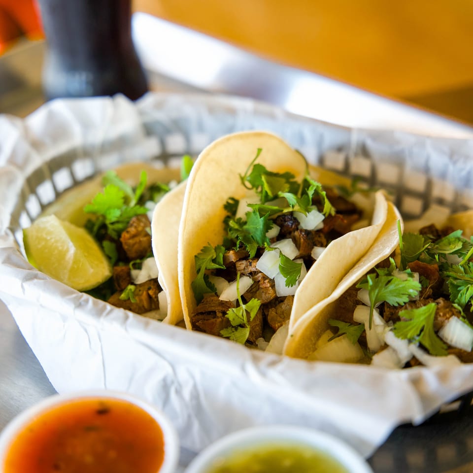 Street-style tacos filled with grilled meat, onions, and cilantro, served with lime wedges, red and green salsa, and a soft drink.