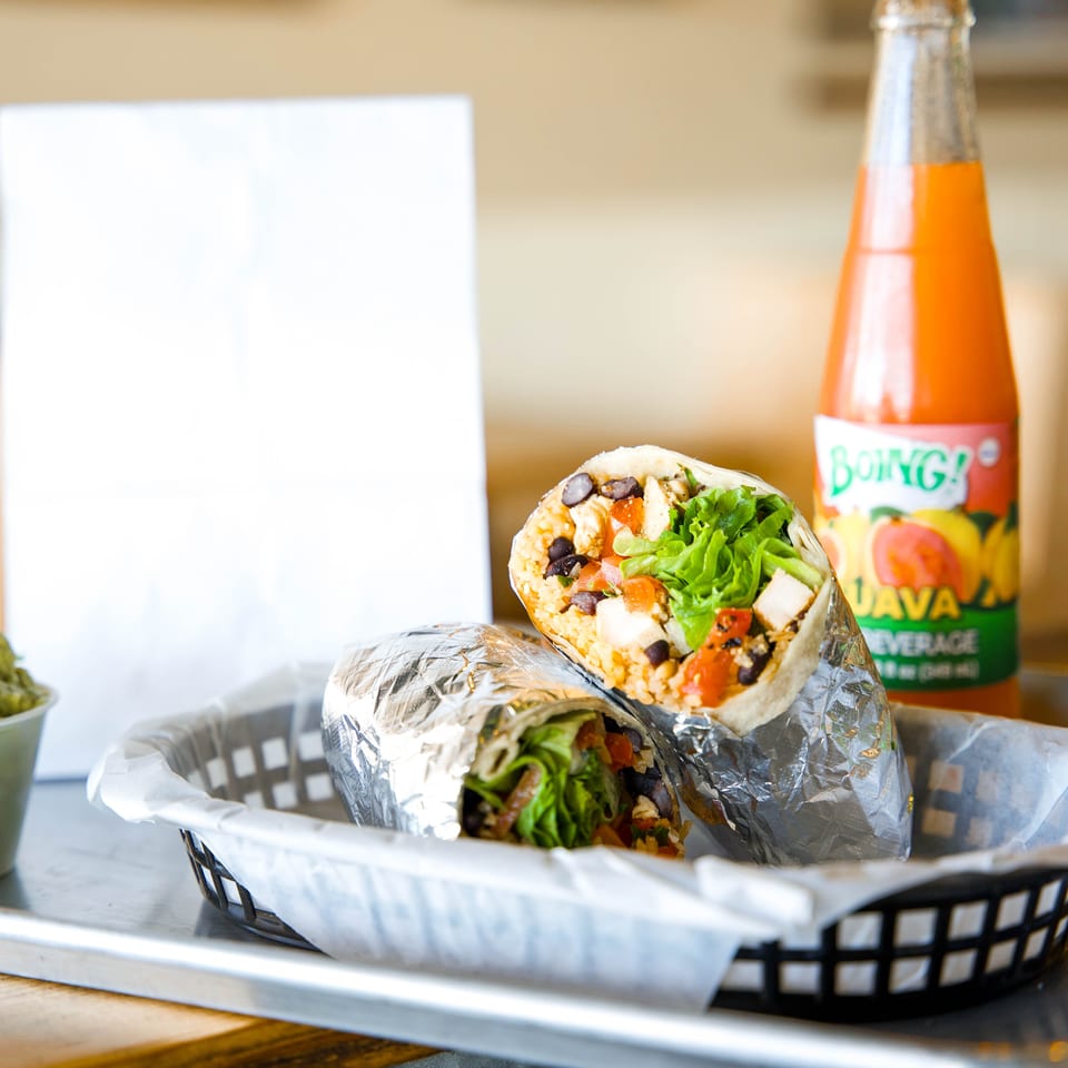 Foil-wrapped burrito filled with rice, black beans, lettuce, chicken, and tomatoes beside a bottle of orange soda.